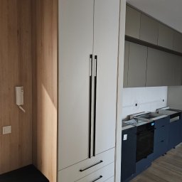 Meble roberto - Nowoczesna zabudowa kuchenna z wysoką szafą w odcieniach bieli i drewna, z czarnymi, minimalistycznymi uchwytami. W tle widoczne szare szafki i przygotowane miejsce na sprzęt.