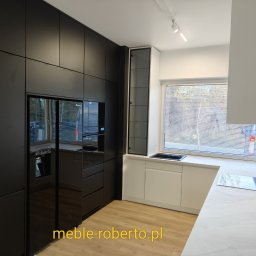 Meble roberto - Nowoczesna kuchnia na wymiar z czarnymi frontami i zabudowaną lodówką, uzupełniona białymi szafkami pod oknem i blatem imitującym marmur. Minimalistyczny design.