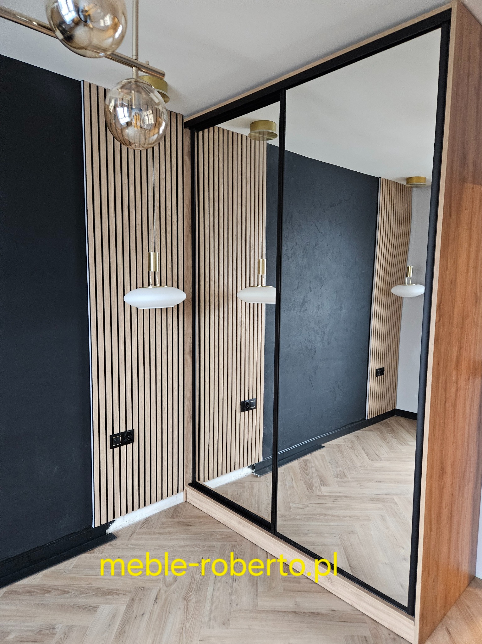 Nowoczesna szafa wnękowa z lustrzanymi drzwiami przesuwnymi, czarne obramowanie, drewniane panele ścienne i designerska lampa sufitowa. Parkiet w jodełkę na podłodze.
