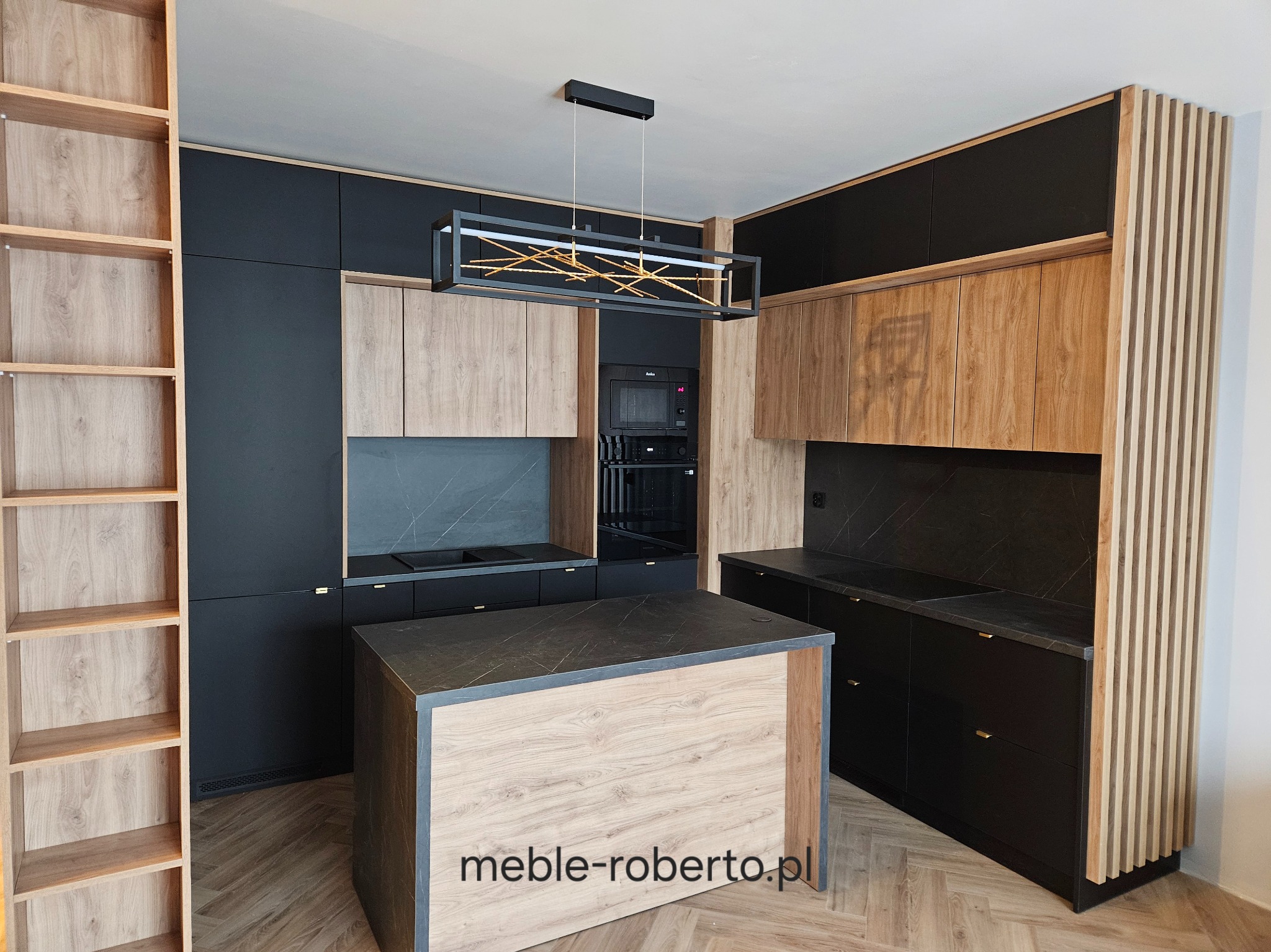 Nowoczesna kuchnia z wyspą: czarne fronty, drewniane akcenty, marmurowy blat. Oświetlenie LED w geometrycznej lampie. Widoczny regał z jasnego drewna i zabudowa piekarnika.