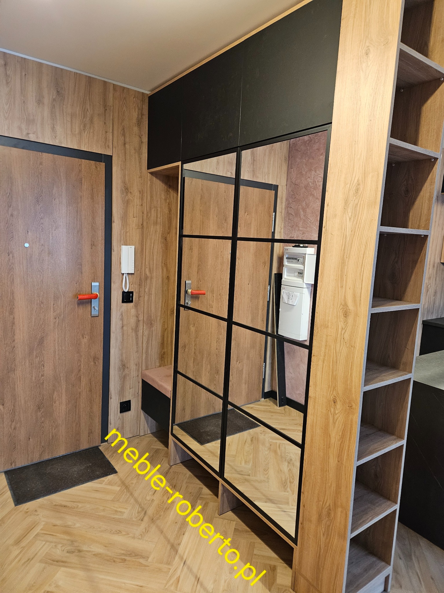 Funkcjonalna zabudowa przedpokoju z lustrzaną szafą w czarnej ramie, drewnianymi panelami i otwartym regałem. Widoczny wieszak i siedzisko.