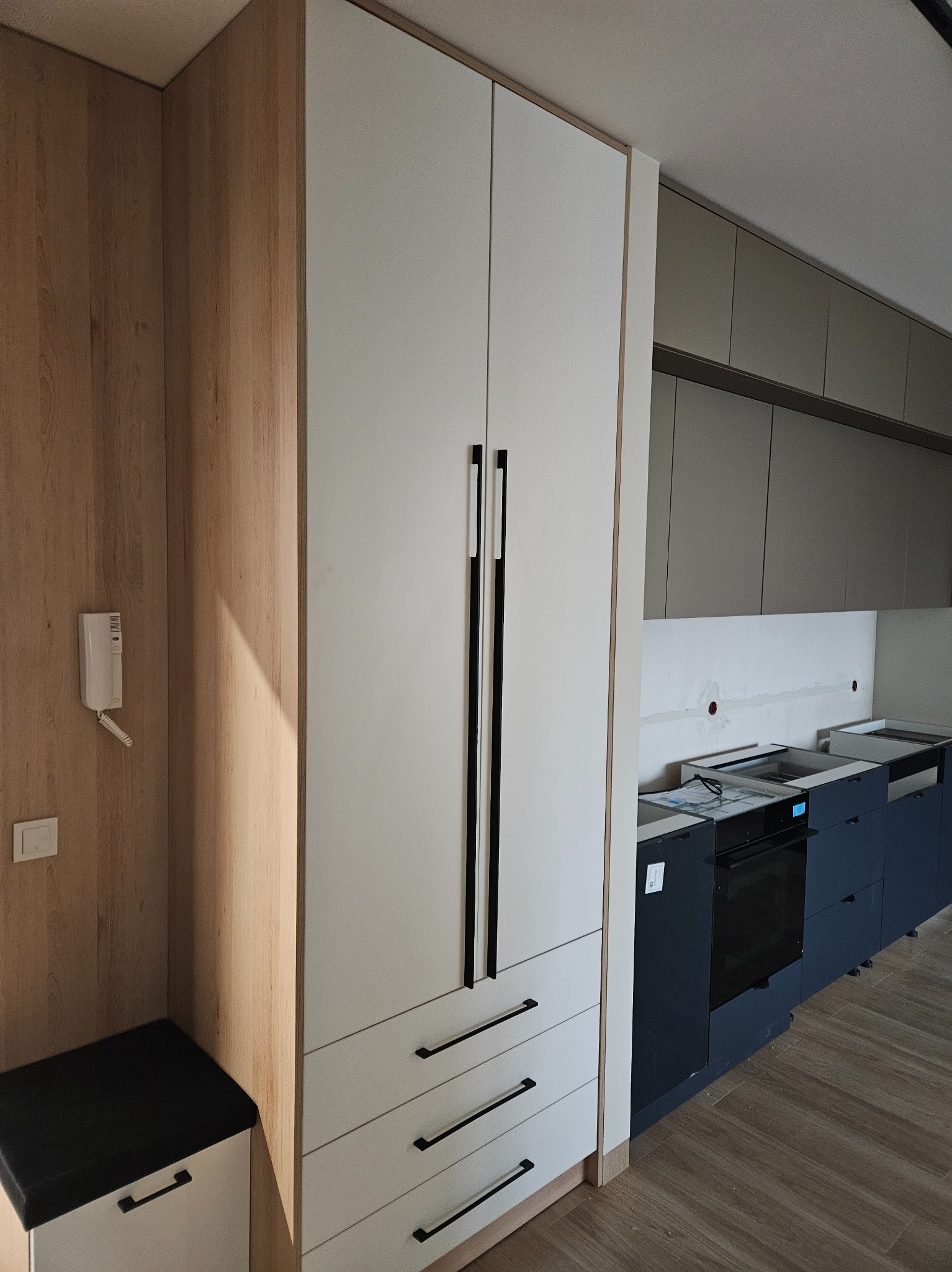 Nowoczesna zabudowa kuchenna z wysoką szafą w odcieniach bieli i drewna, z czarnymi, minimalistycznymi uchwytami. W tle widoczne szare szafki i przygotowane miejsce na sprzęt.