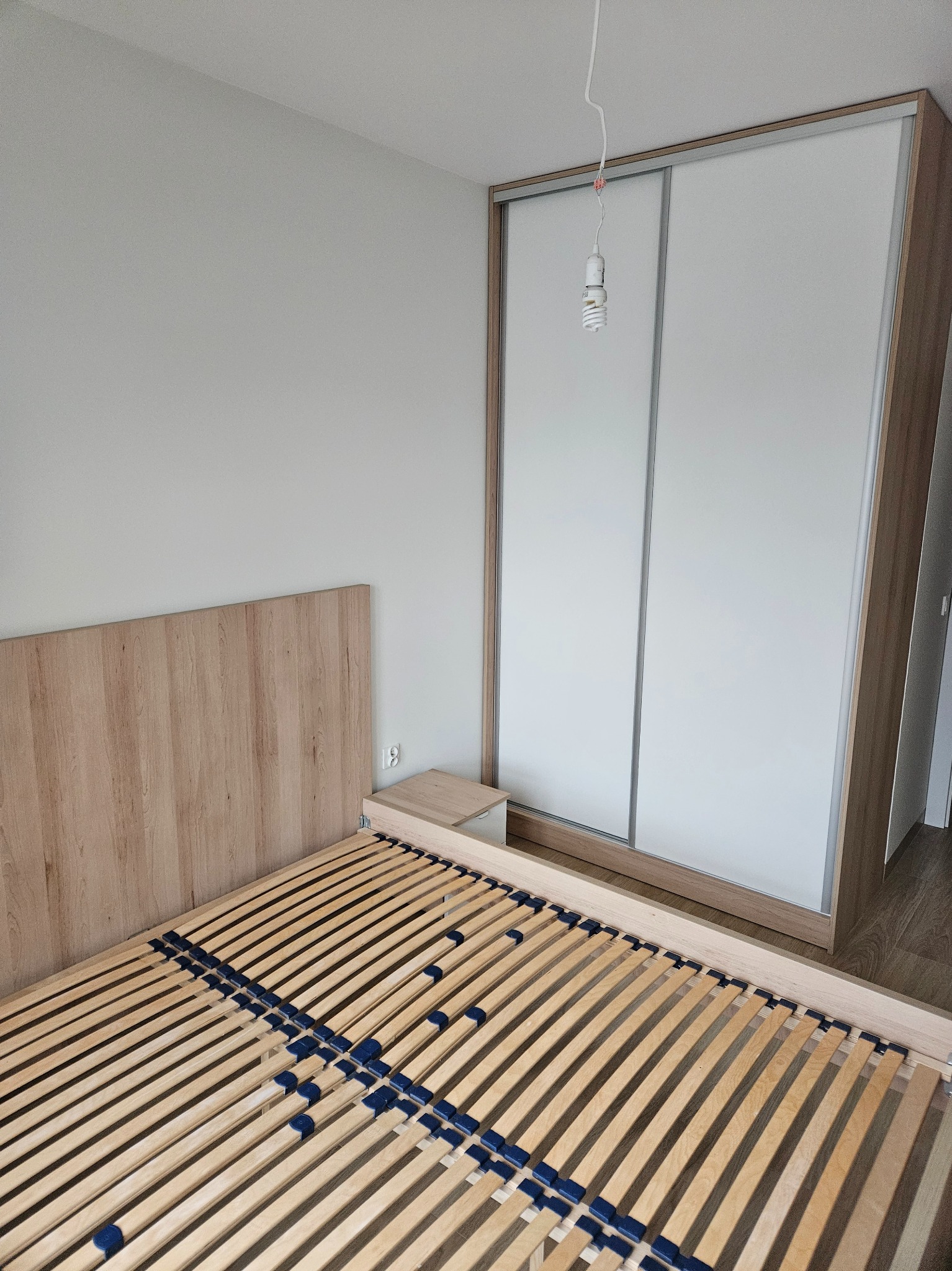 Jasna szafa przesuwna z białymi frontami i drewnianym obramowaniem, obok łóżko z drewnianym zagłówkiem i stelażem. Minimalistyczny design, surowy pokój.