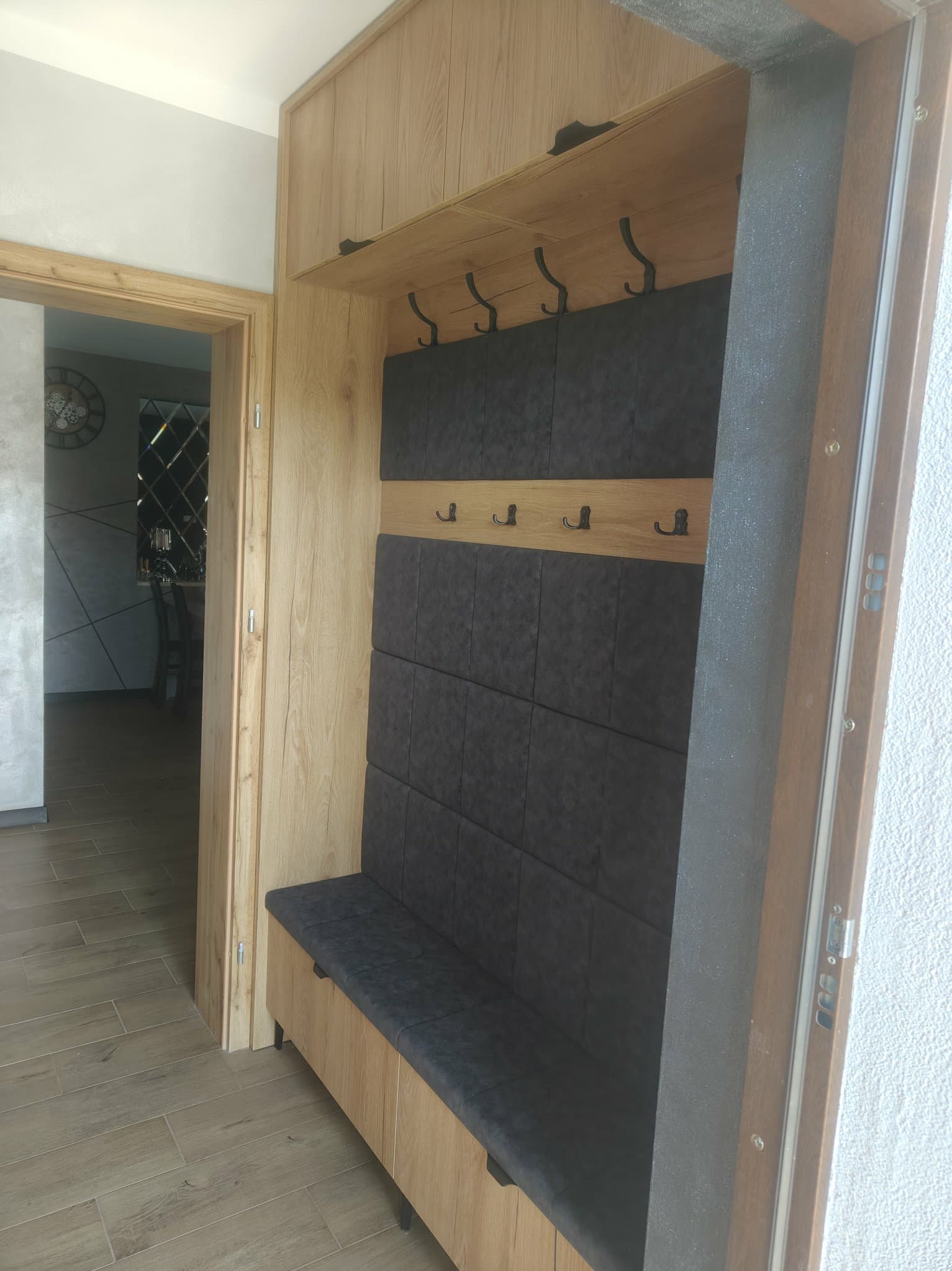 Funkcjonalna zabudowa przedpokoju z siedziskiem i wieszakami, wykończona w drewnie z ciemnoszarą tapicerką. Widok na wnętrze mieszkania przez otwarte drzwi.