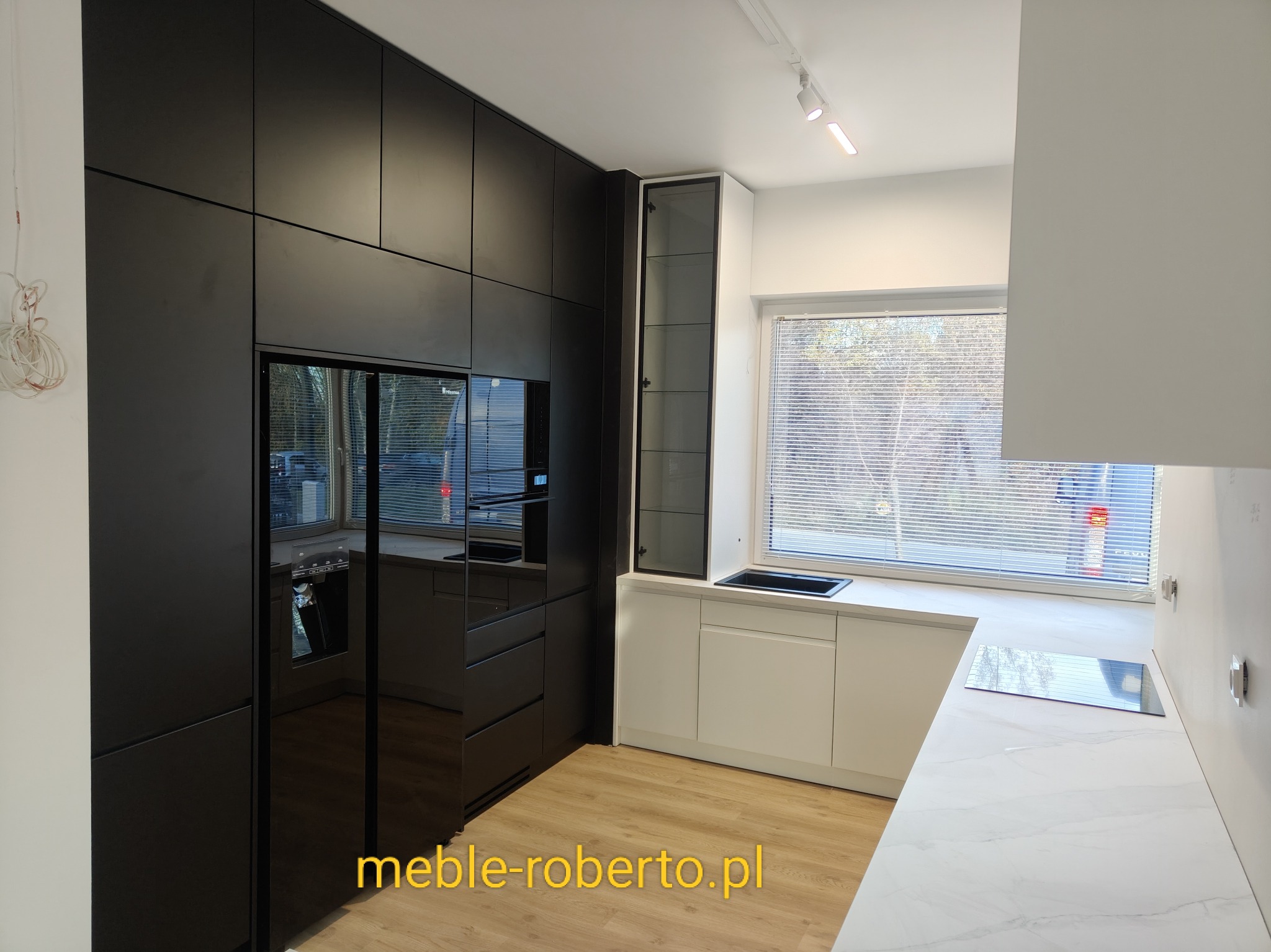 Nowoczesna kuchnia na wymiar z czarnymi frontami i zabudowaną lodówką, uzupełniona białymi szafkami pod oknem i blatem imitującym marmur. Minimalistyczny design.