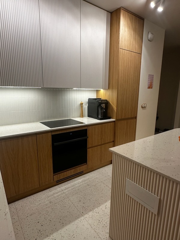 Nowoczesna kuchnia z frezowanymi frontami w odcieniach bieli i drewna, blatem w lastryko i czarnym piekarnikiem. Widoczny ekspres do kawy i wyspa kuchenna.