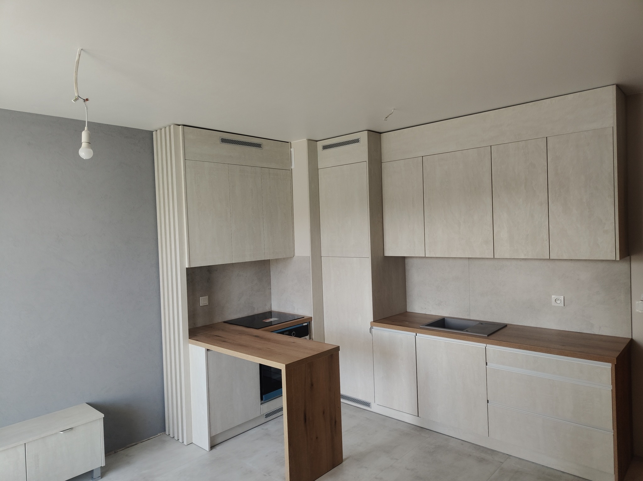 Jasna kuchnia z drewnianym blatem i barkiem, minimalistyczny design, szare ściany, surowy beton na frontach szafek, wisząca żarówka, nowoczesne wykończenie.