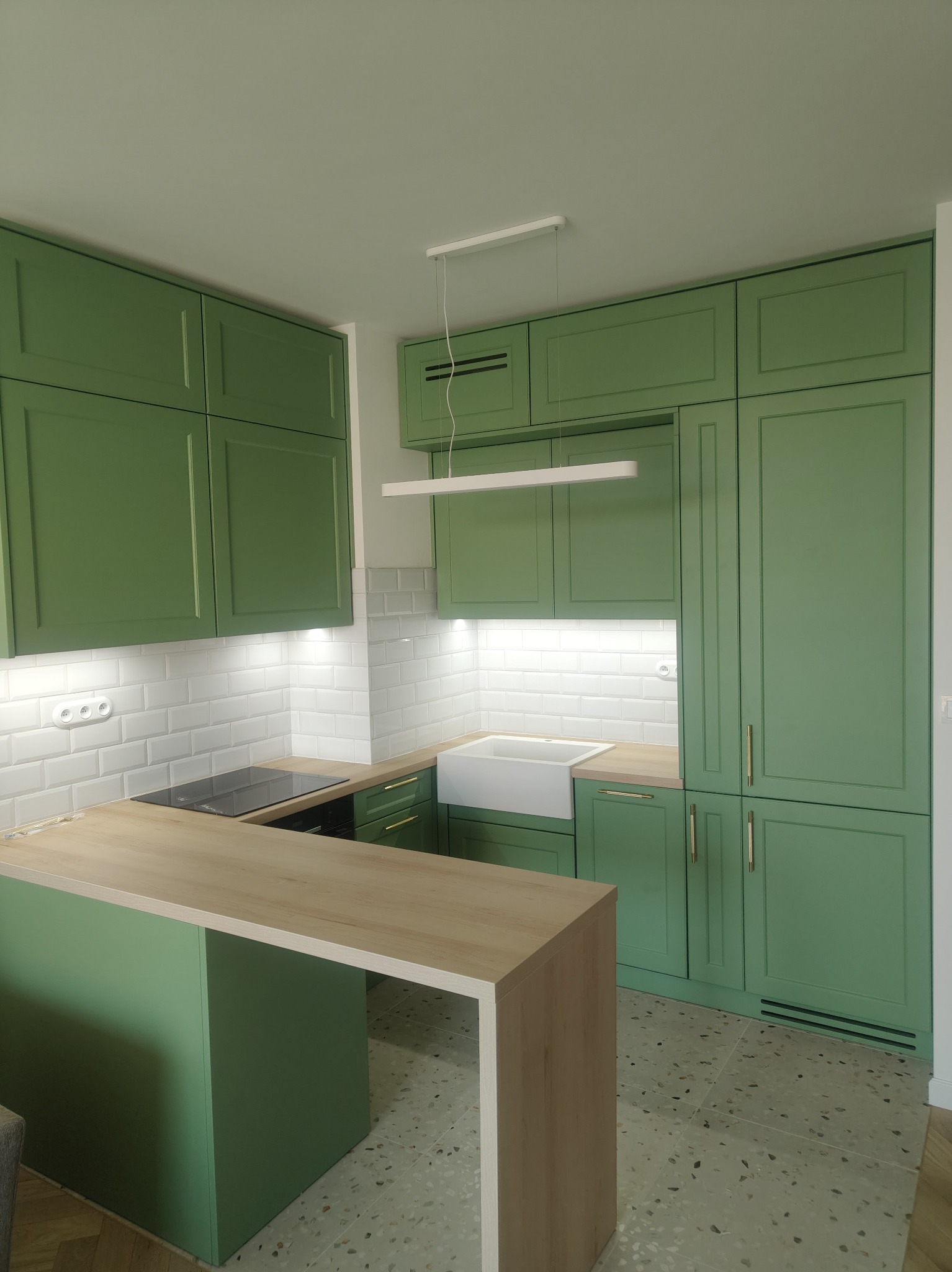 Nowoczesna kuchnia w odcieniach zieleni z drewnianym blatem i białymi płytkami. Uwagę przykuwa minimalistyczna lampa i jasna kolorystyka.