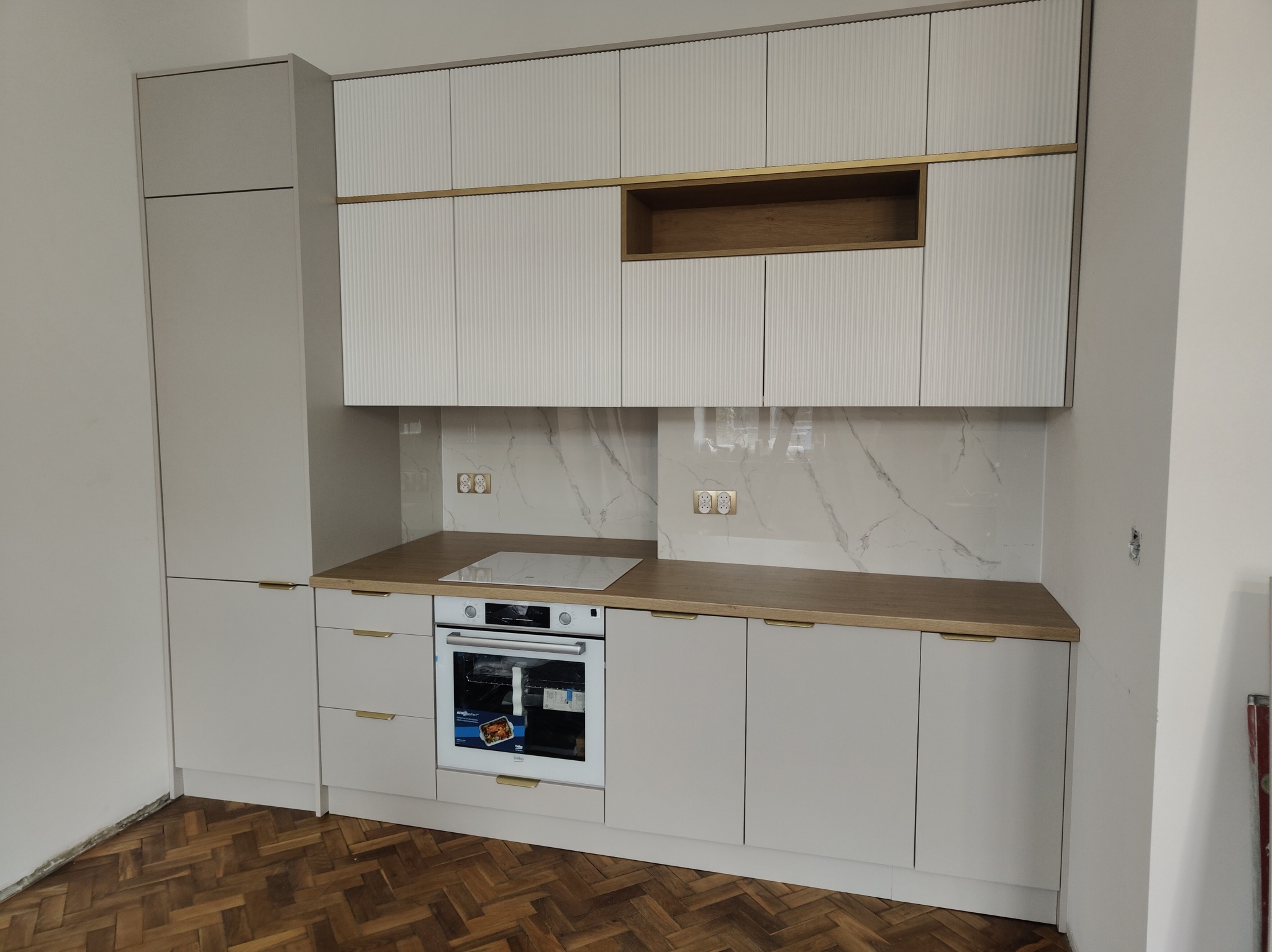 Jasnoszara zabudowa kuchenna z ryflowanymi frontami szafek wiszących, złotymi akcentami i drewnianym blatem. Widoczna płyta indukcyjna i piekarnik.