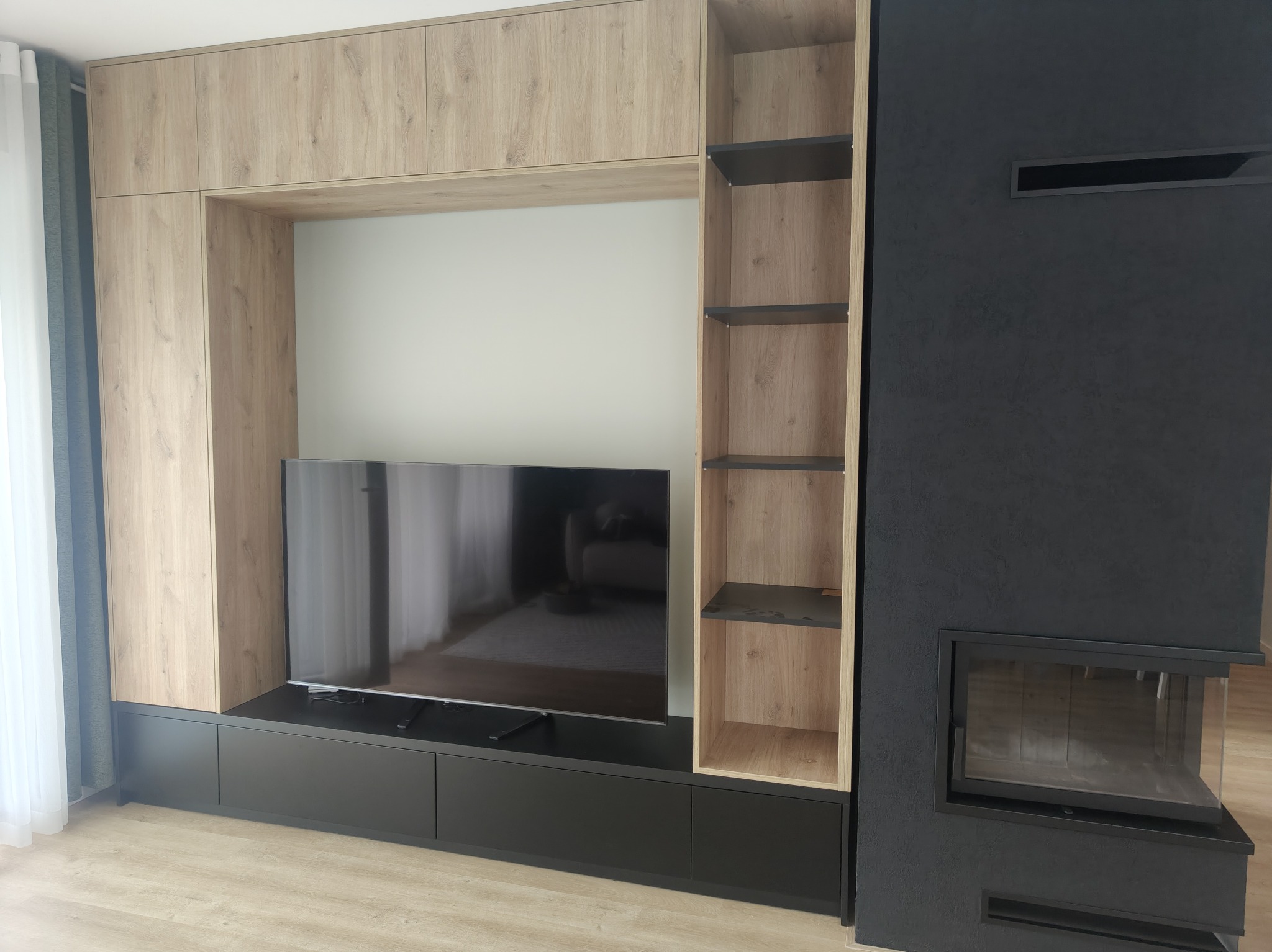 Nowoczesna zabudowa ściany z szafkami, półkami i miejscem na TV, obok kominek w czarnej obudowie. Jasne drewno i czerń tworzą elegancki kontrast.