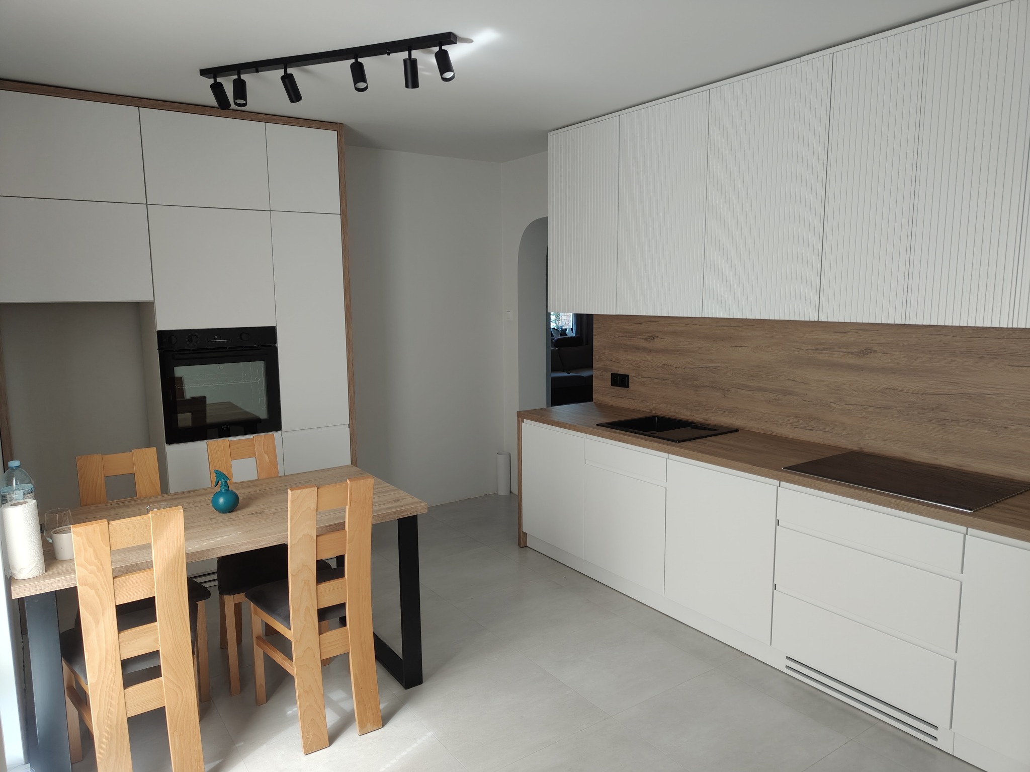 Jasna kuchnia z białymi frontami i drewnianym blatem. Widoczny stół z krzesłami. Minimalistyczny design, oświetlenie punktowe. Ujęcie z perspektywy wnętrza.