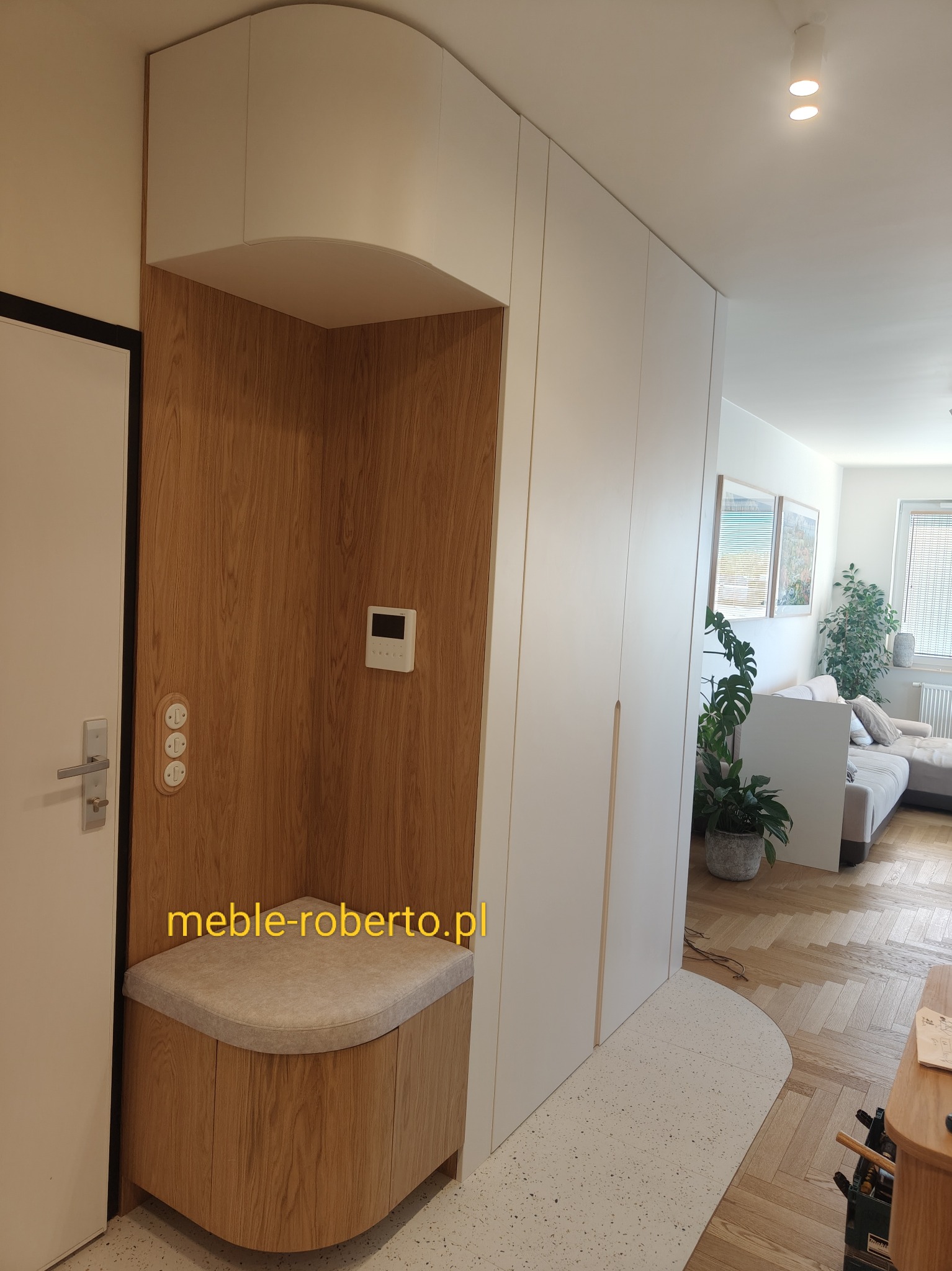 Funkcjonalna zabudowa przedpokoju z siedziskiem i szafą wnękową w kolorze białym i drewnopodobnym. Nowoczesny design, widok na salon z roślinami.