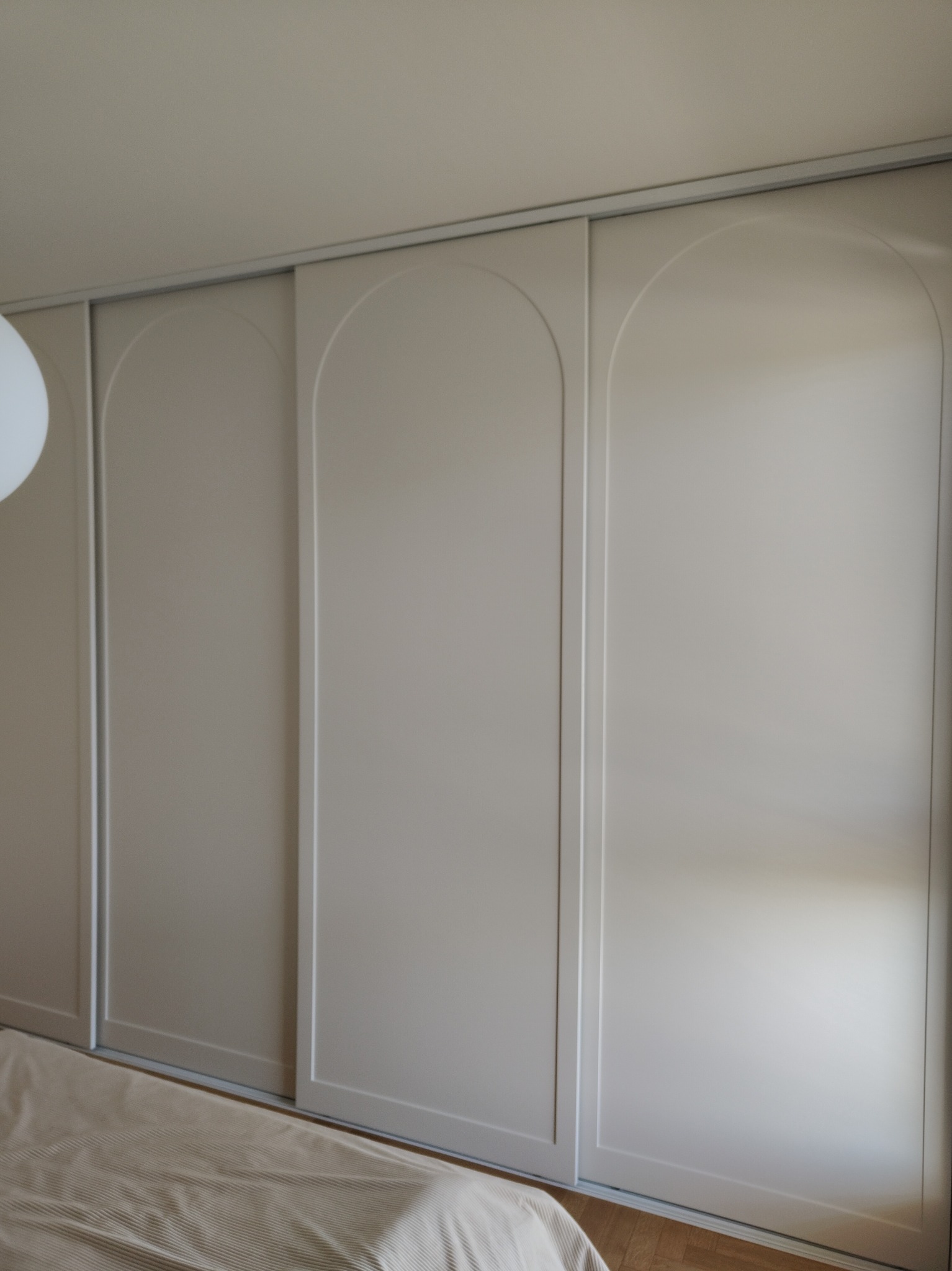 Beżowa szafa wnękowa z przesuwnymi drzwiami o łukowatym wzorze, widoczna częściowo pościel w paski, minimalistyczny design, jasne wnętrze.