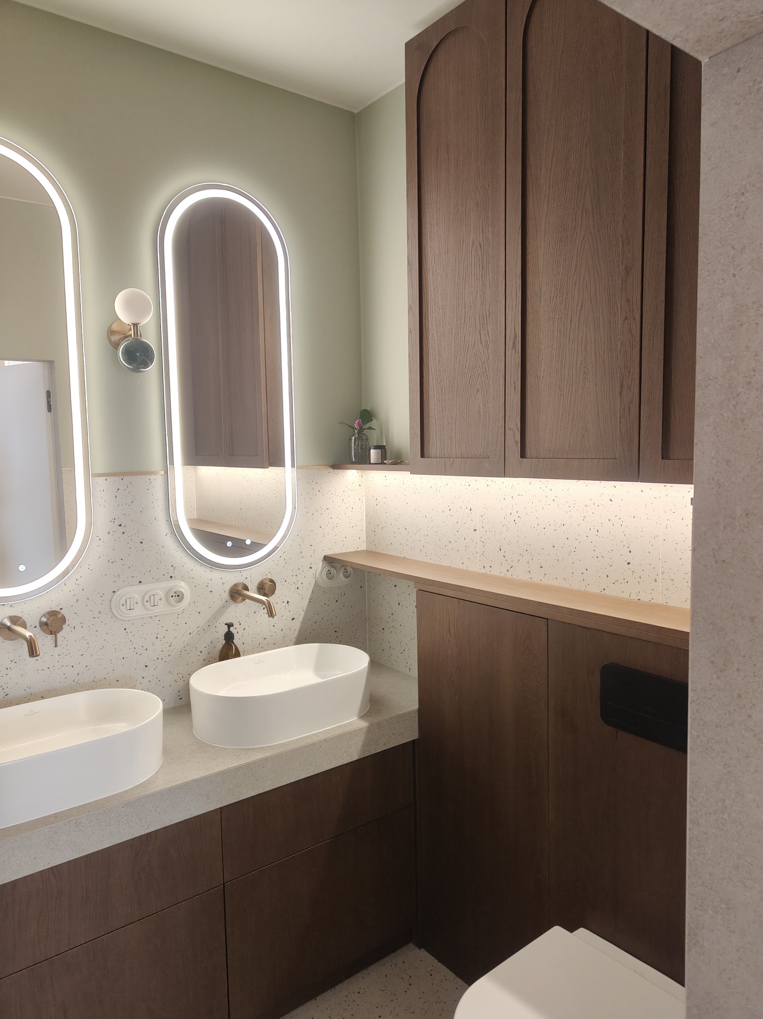 Nowoczesna łazienka z dwoma umywalkami nablatowymi, podświetlanymi lustrami w kształcie łuku i szafkami z ciemnego drewna. Ściany wykończone płytkami lastryko.