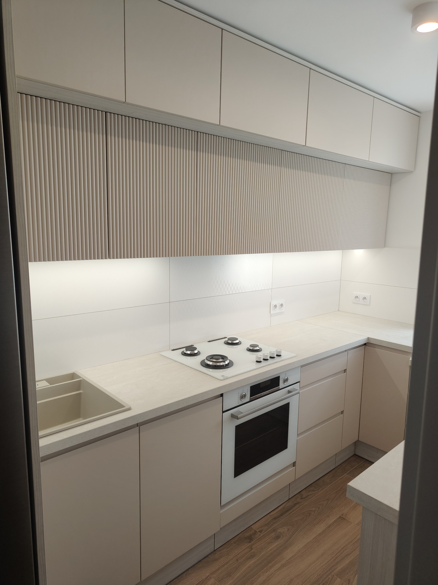 Jasna kuchnia z frezowanymi frontami szafek górnych, białym blatem i sprzętem AGD. Ujęcie z perspektywy wejścia, widoczna podłoga z paneli.