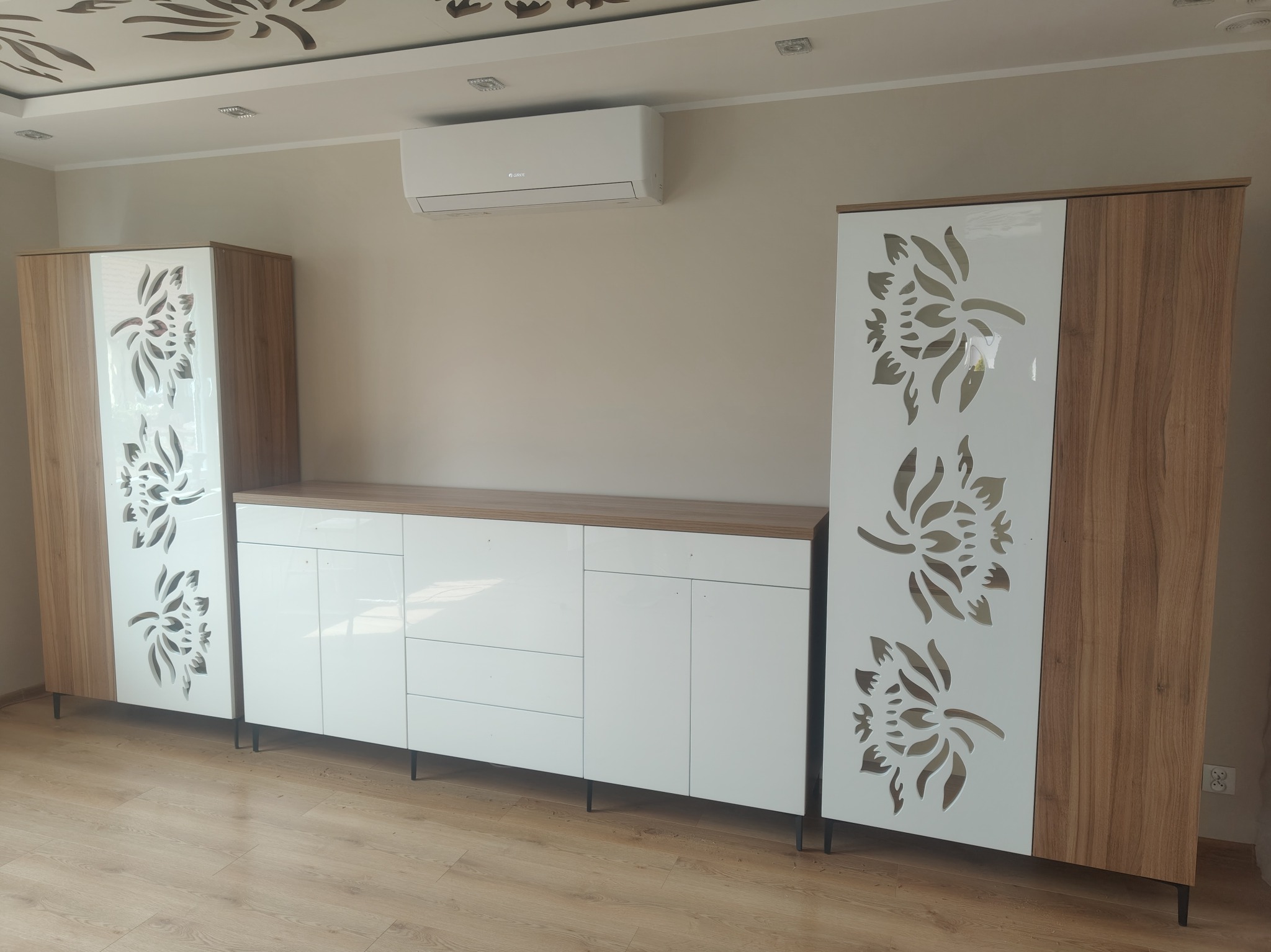 Nowoczesny zestaw mebli: biała komoda z drewnianym blatem i dwie szafy z dekoracyjnymi, ażurowymi frontami w kwiatowy wzór. Całość dopełnia klimatyzacja na ścianie.