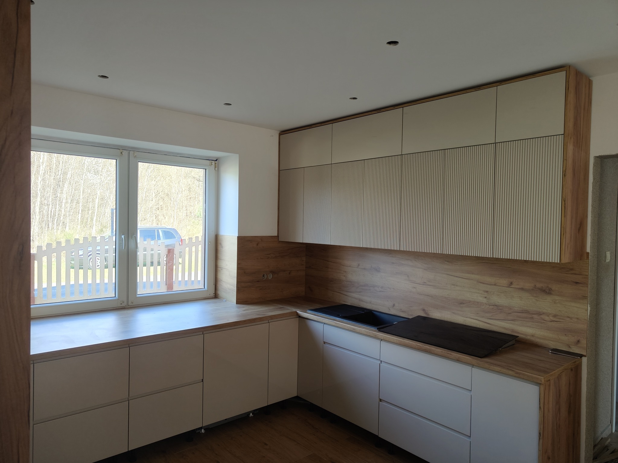 Jasna kuchnia z drewnianymi akcentami: fronty szafek w odcieniach beżu i drewna, blat z płyty drewnopodobnej, okno z widokiem na zieleń. Nowoczesny design.