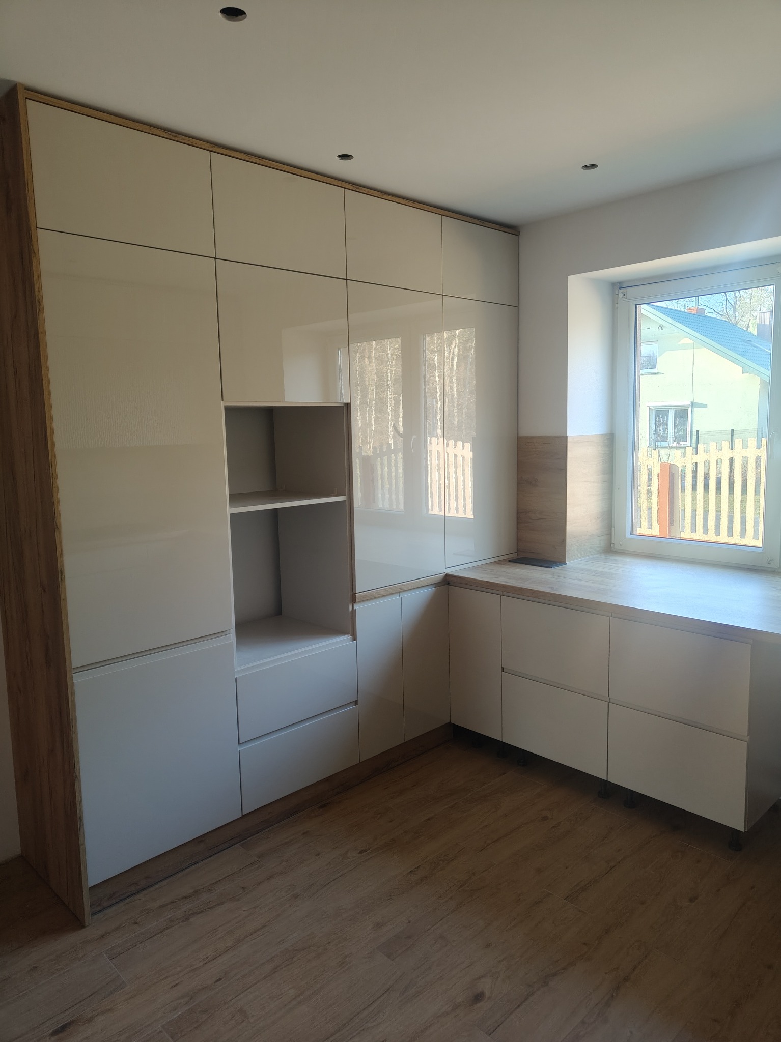 Jasna zabudowa kuchenna z elementami drewnopodobnymi, obejmująca szafki, półki i blat pod oknem, na tle drewnianej podłogi i białych ścian.