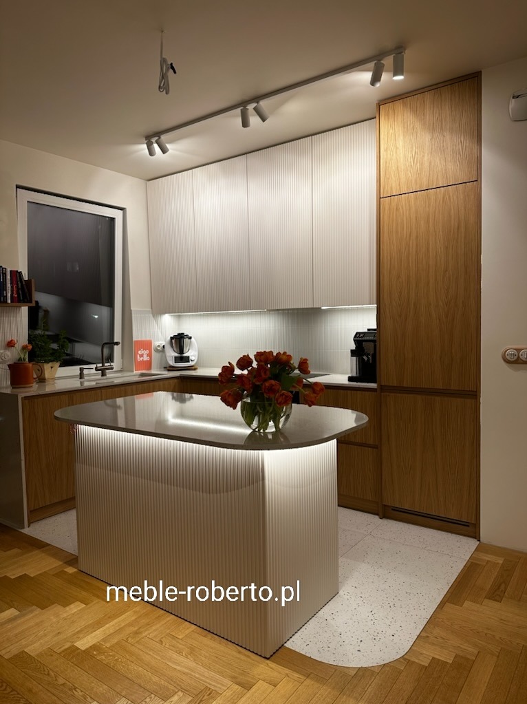 Nowoczesna kuchnia z wyspą, białe ryflowane fronty szafek górnych i wyspy, drewniane szafki dolne, oświetlenie LED pod szafkami i na wyspie, bukiet czerwonych tulipanów.