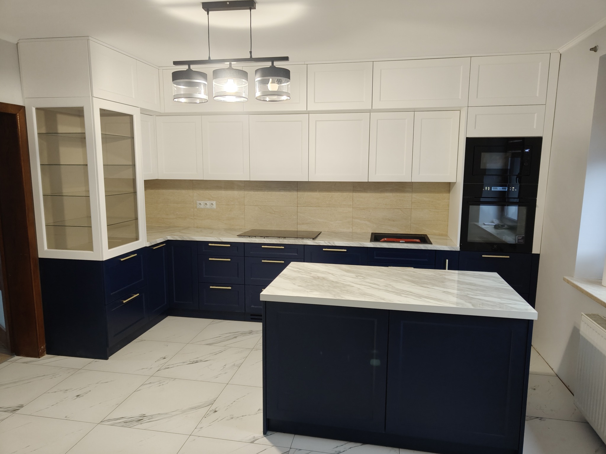 Nowoczesna kuchnia z wyspą: granatowe szafki dolne z złotymi uchwytami, białe szafki górne, marmurowy blat i podłoga. Oświetlenie sufitowe.