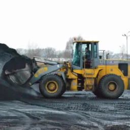 Żółta ładowarka ZL50G nabiera węgiel z pryzmy, widoczne ślady opon na ziemi, w tle drzewa i słupy oświetleniowe.