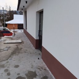 LS-BUD - Biała elewacja budynku z brązowym cokołem i czarnymi lampami sufitowymi. Widoczny fragment chodnika i otoczenie budynku.