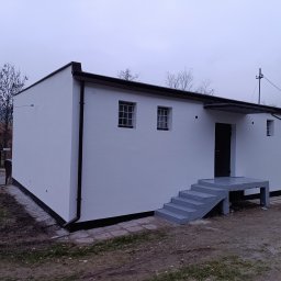 LS-BUD - Biały, parterowy budynek z ciemnymi rynnami i schodami wejściowymi, otoczony zielenią i drzewami, sfotografowany w pochmurny dzień. Widoczne okna z kratami.