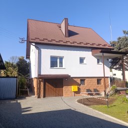 LS-BUD - Dwukondygnacyjny dom z brązowym dachem i białymi ścianami, z dekoracyjną elewacją na parterze. Widoczny podjazd z kostki brukowej i fragment ogrodu.