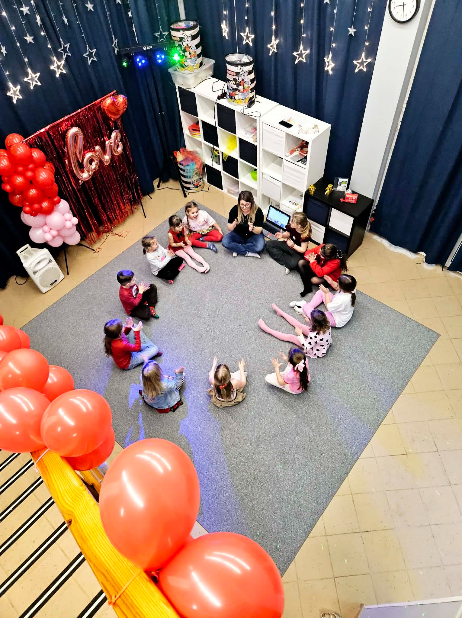 Animator prowadzi zajęcia dla dzieci siedzących w kręgu na dywanie. Dekoracje: balony, kurtyna z gwiazdami, napis LOVE. Widok z góry, oświetlenie dyskotekowe.