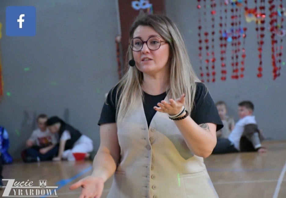 Animator w beżowej kamizelce, z mikrofonem, gestykuluje na tle sali z dziećmi i dekoracjami. Widoczny logotyp firmy w lewym dolnym rogu.