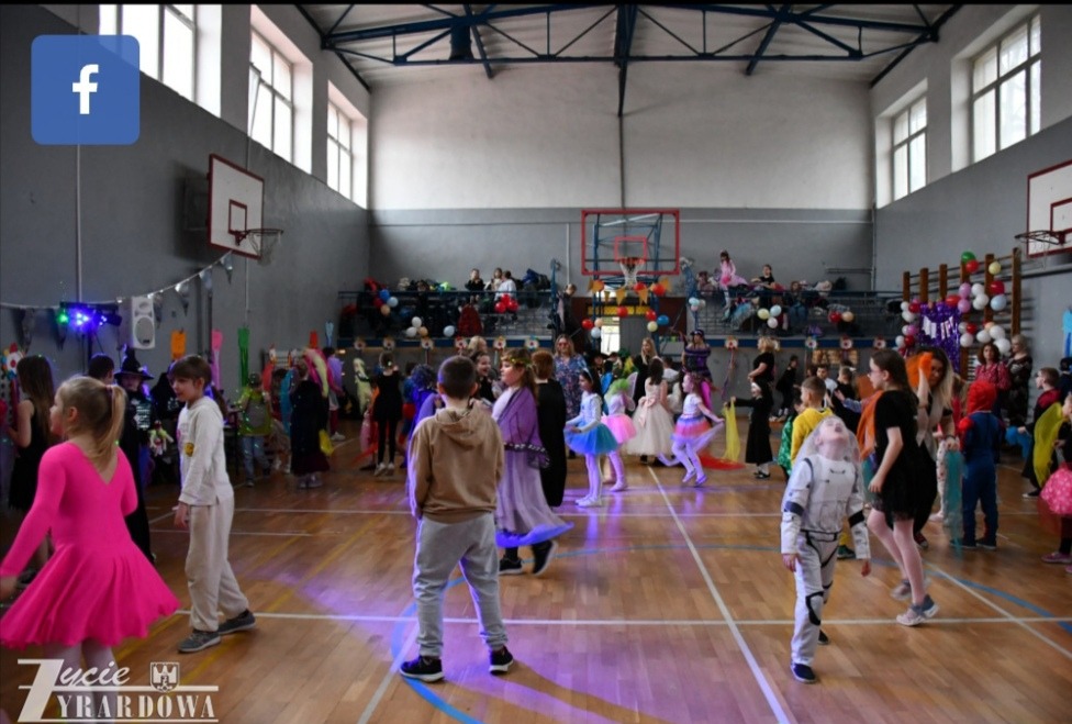 Sala gimnastyczna udekorowana na bal przebierańców. Dzieci w kostiumach tańczą, a rodzice obserwują z trybun. Dekoracje z balonów i światełek.