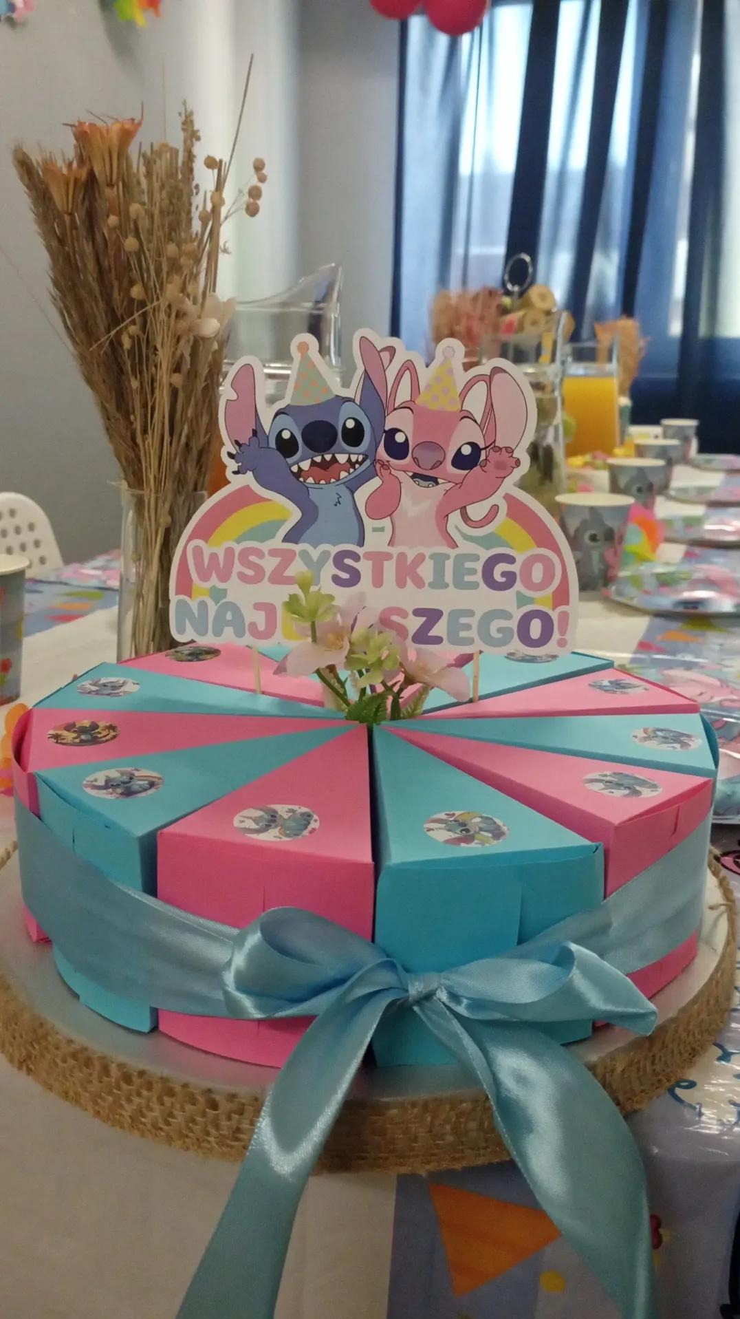 Tort z motywem Lilo i Stitch, podzielony na porcje w pudełkach, przewiązany błękitną wstążką. Dekoracja stołu urodzinowego z napisem 'Wszystkiego Najlepszego!'.