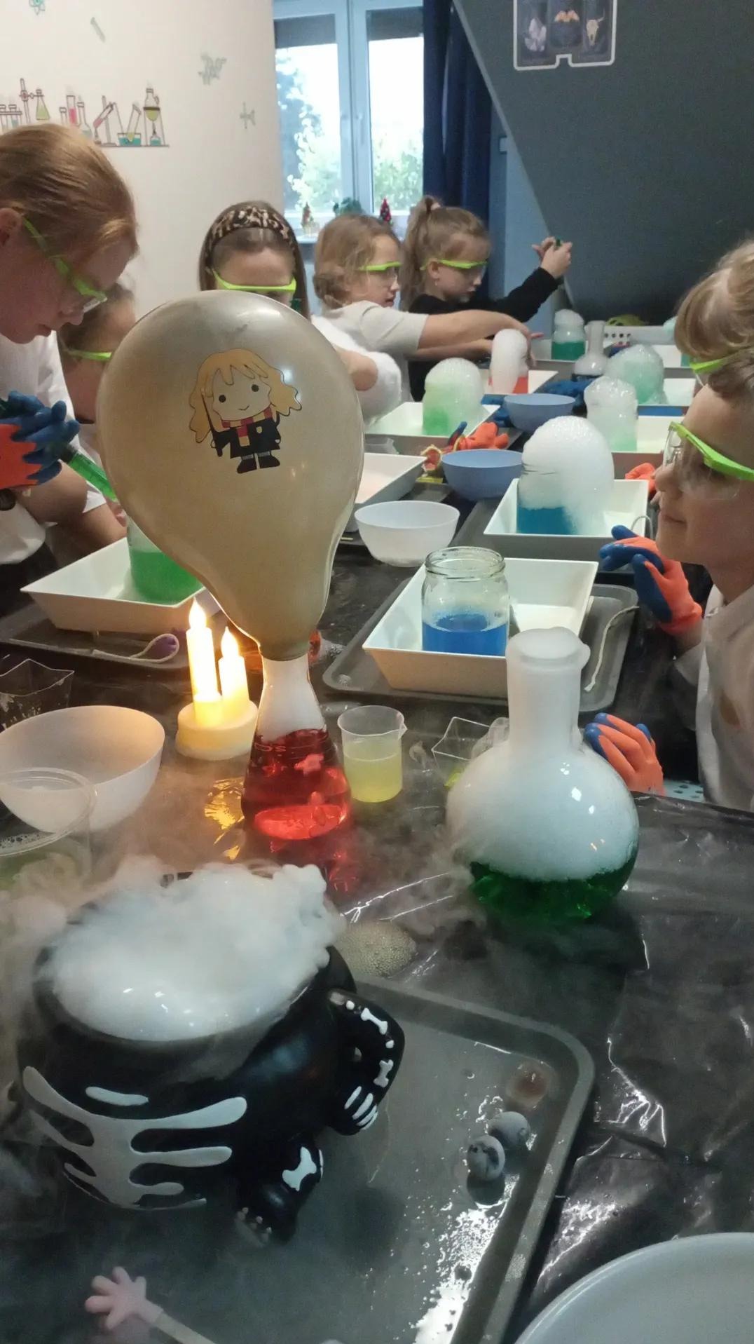 Dzieci w okularach ochronnych uczestniczą w eksperymentach chemicznych podczas imprezy urodzinowej z motywem Harry'ego Pottera, z użyciem dymiących mikstur i balonów.