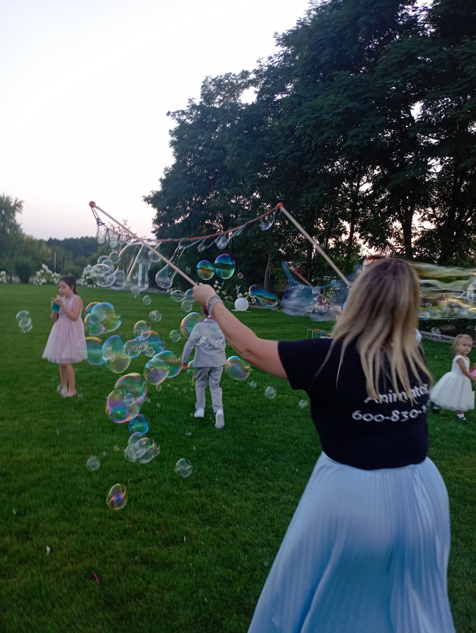 Animator dmucha duże bańki mydlane na zielonym trawniku podczas imprezy plenerowej dla dzieci. Widoczne dzieci w strojach wizytowych i sprzęt do robienia baniek.