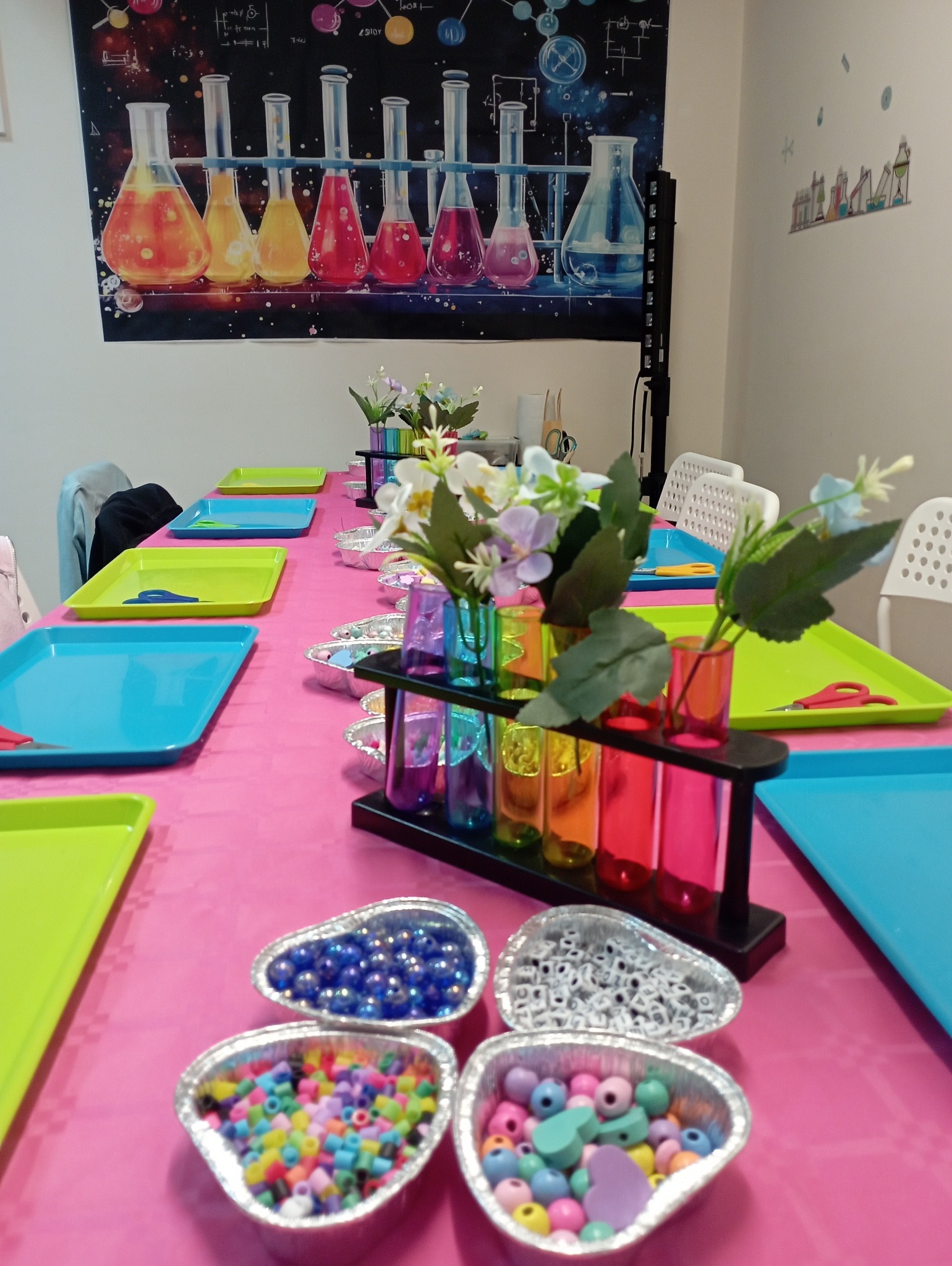 Stół z kolorowymi dekoracjami i naczyniami laboratoryjnymi, przygotowany do kreatywnych zajęć dla dzieci, z tęczowymi kwiatami w probówkach i różowym obrusem.