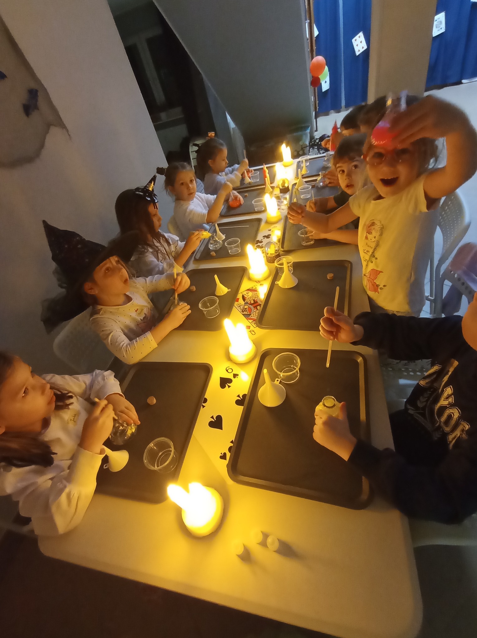 Dzieci przy stole dekorowanym kartami, uczestniczące w warsztatach z elementami magii, oświetlone świecami LED, jedna z dziewczynek trzyma w ręku czerwoną kulkę.