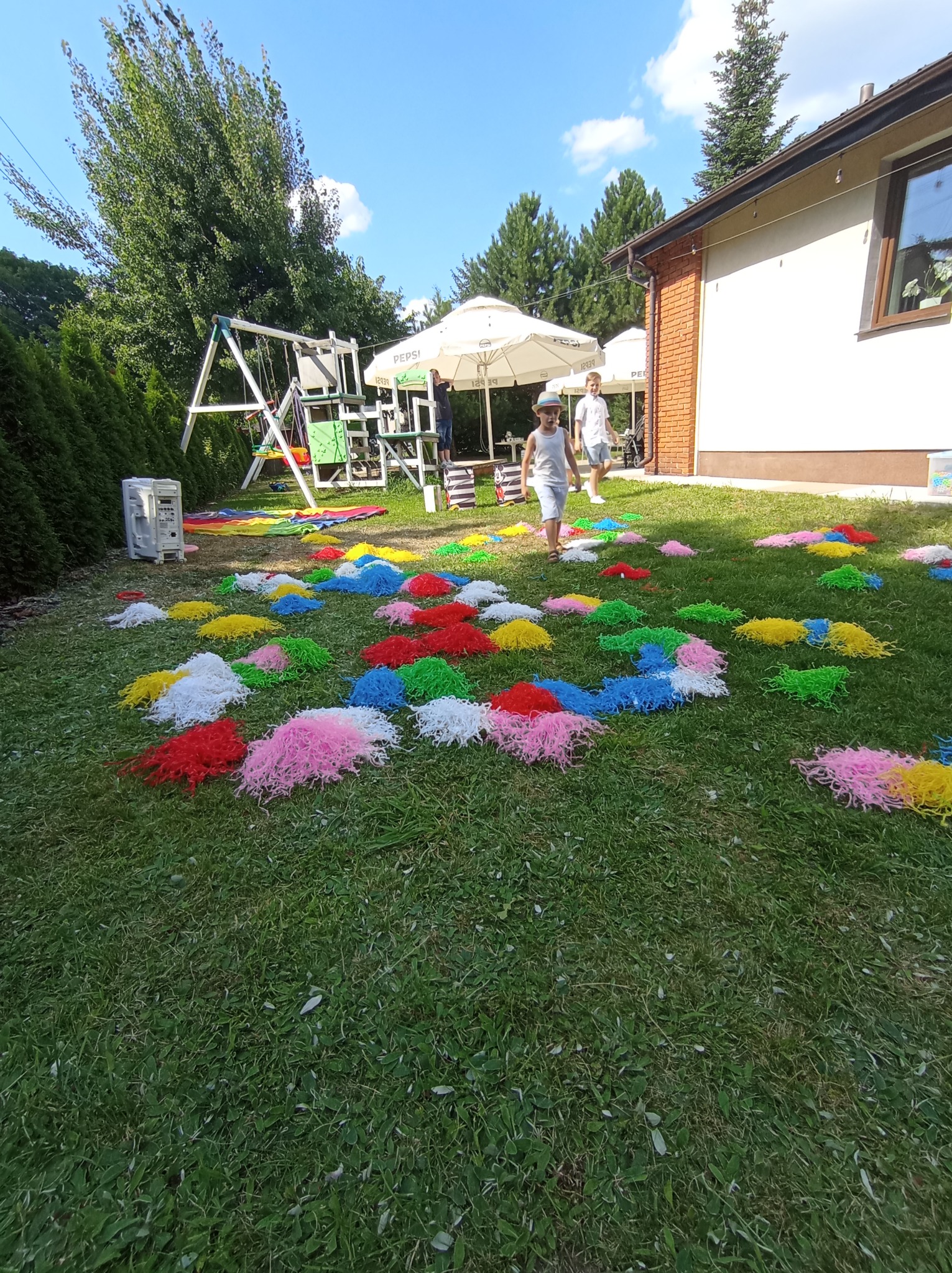 Kolorowe pompony na trawie podczas animacji dla dzieci w plenerze. W tle huśtawka, parasole i budynek. Dziecko w kapeluszu idzie po trawie.