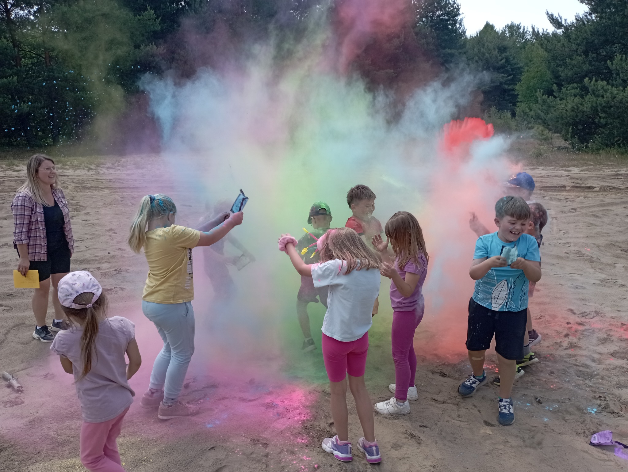 Grupa dzieci i dorosła kobieta w plenerze, rzucają kolorowe proszki holi, tworząc chmurę barw. Zabawa na piaszczystym terenie, w tle drzewa.