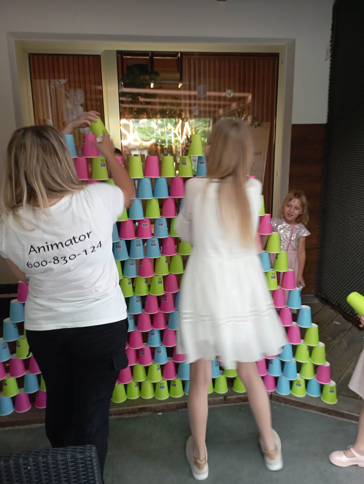 Animator buduje piramidę z kolorowych kubków podczas imprezy dla dzieci. Widoczna dziewczynka w sukience i numer telefonu na koszulce animatora.