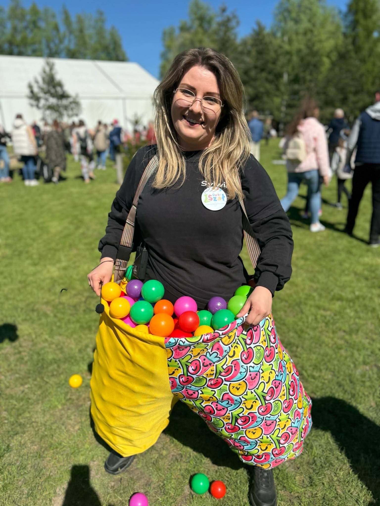 Uśmiechnięta animatorka w czarnej bluzie z kolorowymi piłkami w worku na trawie, w tle osoby i biały namiot. Słoneczny dzień, impreza plenerowa.