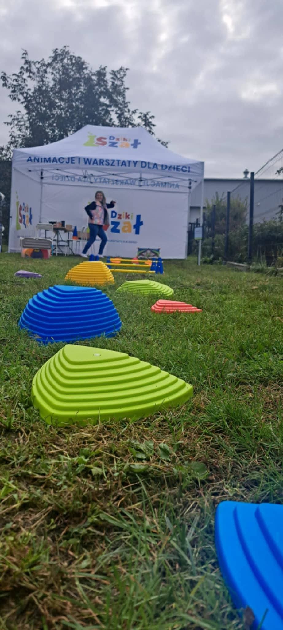 Dziewczynka na torze przeszkód z kolorowych elementów, w tle namiot z napisem 'Animacje i warsztaty dla dzieci'. Aktywna zabawa na świeżym powietrzu.