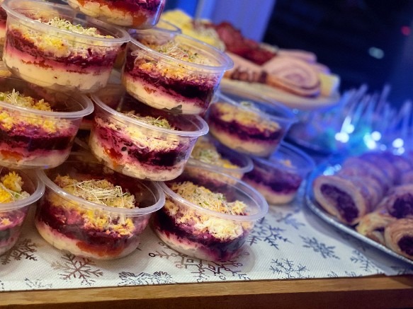 Piramida z porcjowanych sałatek warstwowych w plastikowych pojemnikach, dekorowanych kiełkami, na stole z motywem śnieżynek. W tle wędliny i paszteciki.