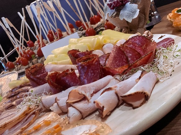 Bogato zastawiony stół z przekąskami: wędliny, sery, koreczki z pomidorkami koktajlowymi. Dekoracja z kwiatów i ziół. Idealne na przyjęcie firmowe.