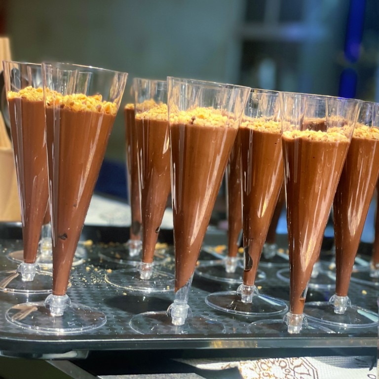 Deser czekoladowy w przezroczystych, stożkowych naczyniach, posypany kruszonką, ustawiony na czarnej tacy. Widoczne refleksy światła na szkle.