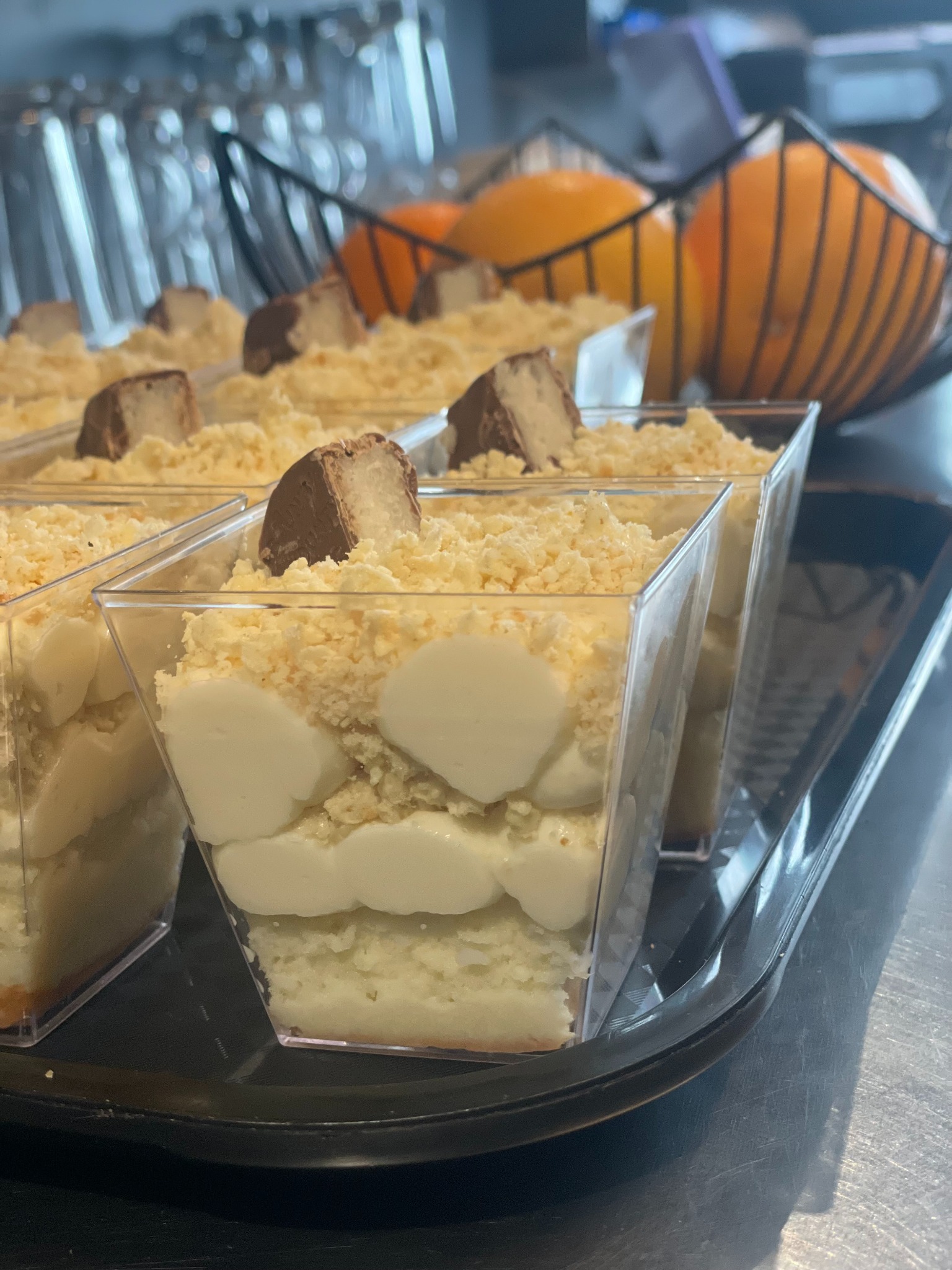 Deser w przezroczystych pojemnikach z kremem, posypką i kawałkiem czekolady na czarnej tacy. W tle koszyk z pomarańczami.