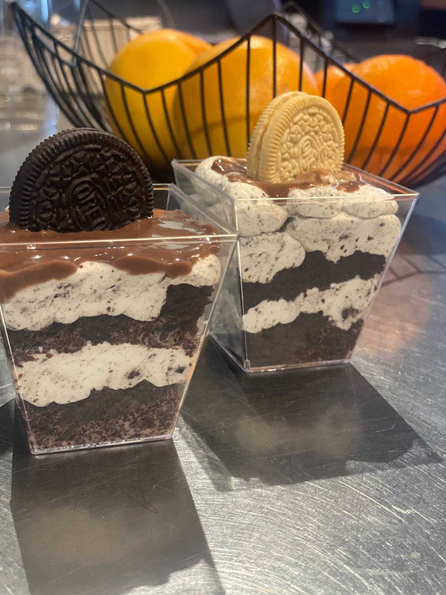 Dwa desery Oreo w przezroczystych pojemnikach, z czarnym i złotym ciasteczkiem na wierzchu, na metalowym blacie, z koszem pomarańczy w tle.