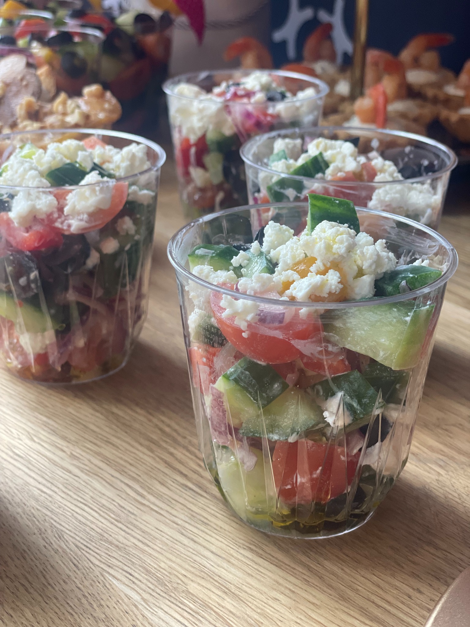 Sałatka grecka w przezroczystych kubeczkach na drewnianym stole. Widoczne pomidory, ogórki, oliwki i ser feta. Catering na imprezę.