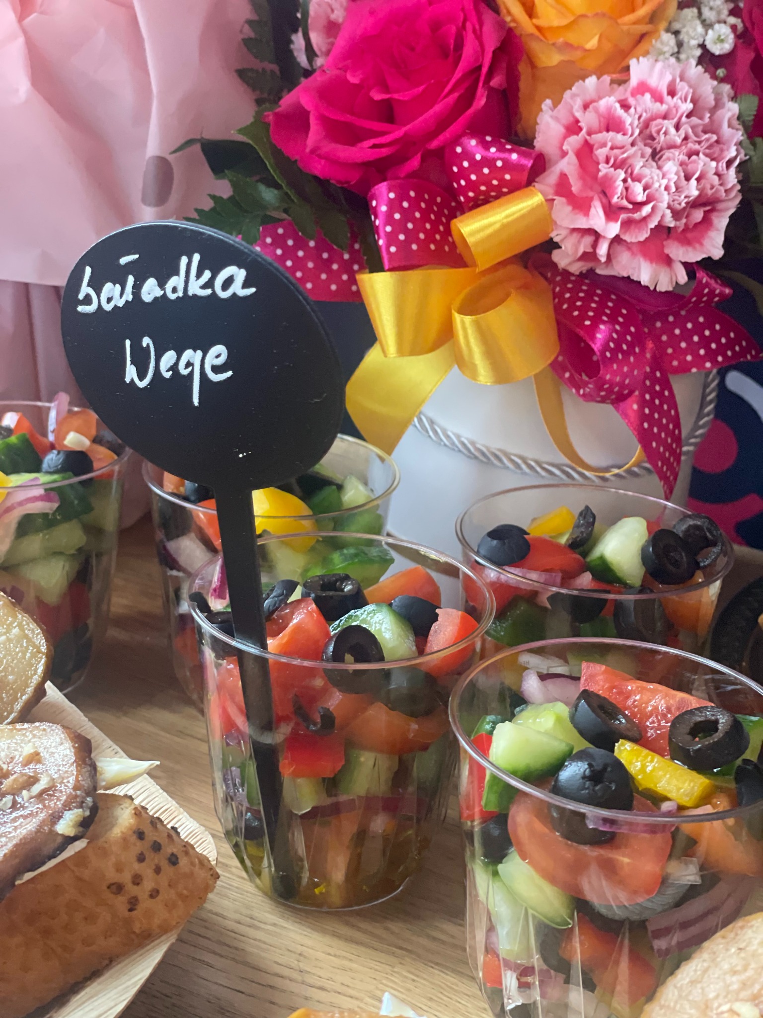 Szklanki z sałatką wege, dekorowane kwiatami i wstążkami. Na pierwszym planie tabliczka z napisem 'Sałatka Wege'. Catering na przyjęcie.
