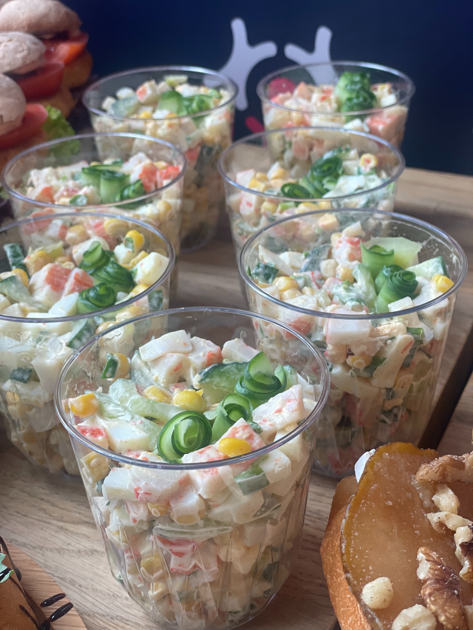 Sałatka krabowa w przezroczystych kubeczkach, dekorowana spiralą z ogórka, na drewnianej desce. Widoczne też kanapki i deser z gruszką.