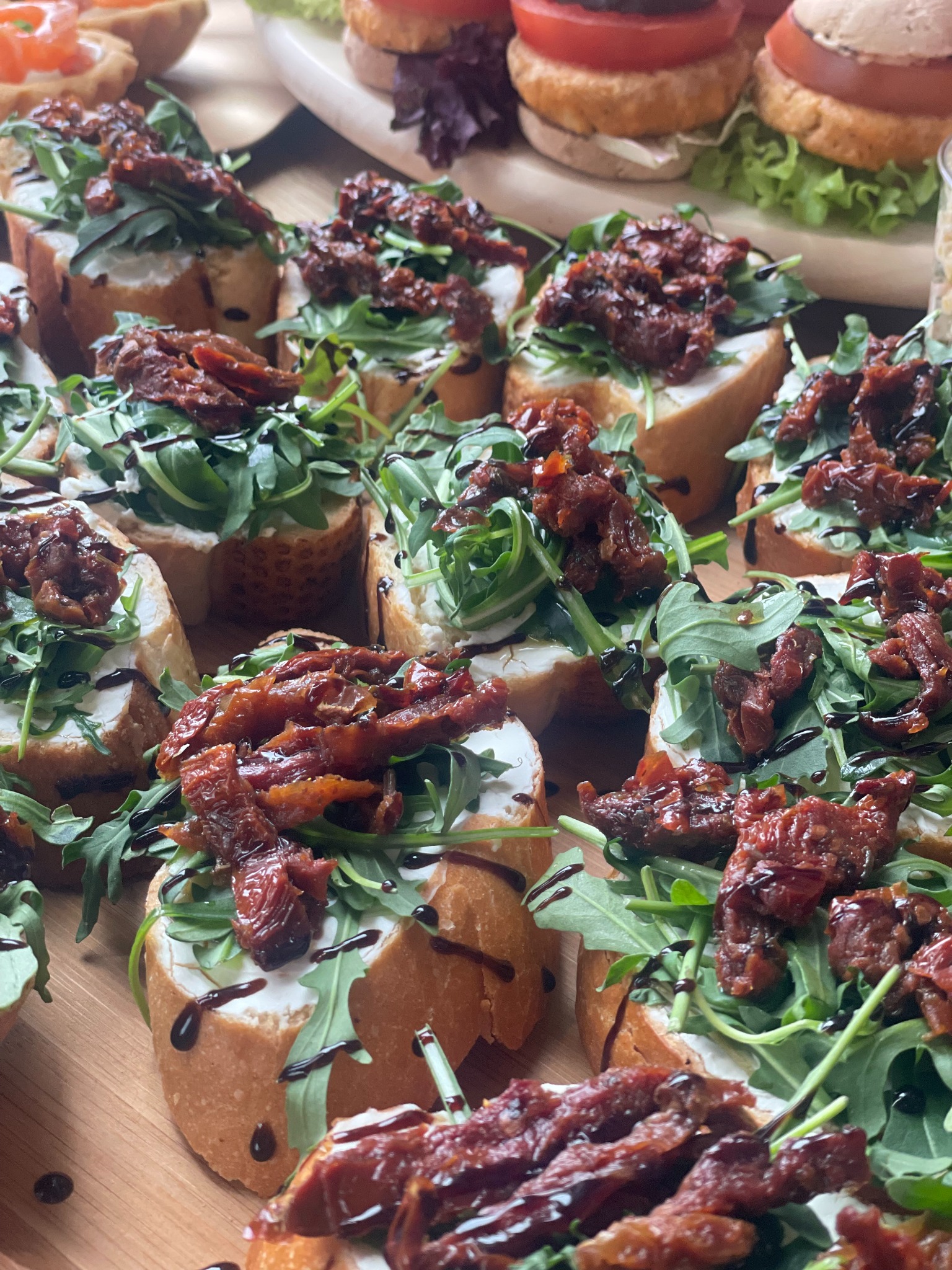 Bruschetta z rukolą, suszonymi pomidorami i kremowym serem, polana balsamicznym sosem. Eleganckie przekąski cateringowe na drewnianej desce.